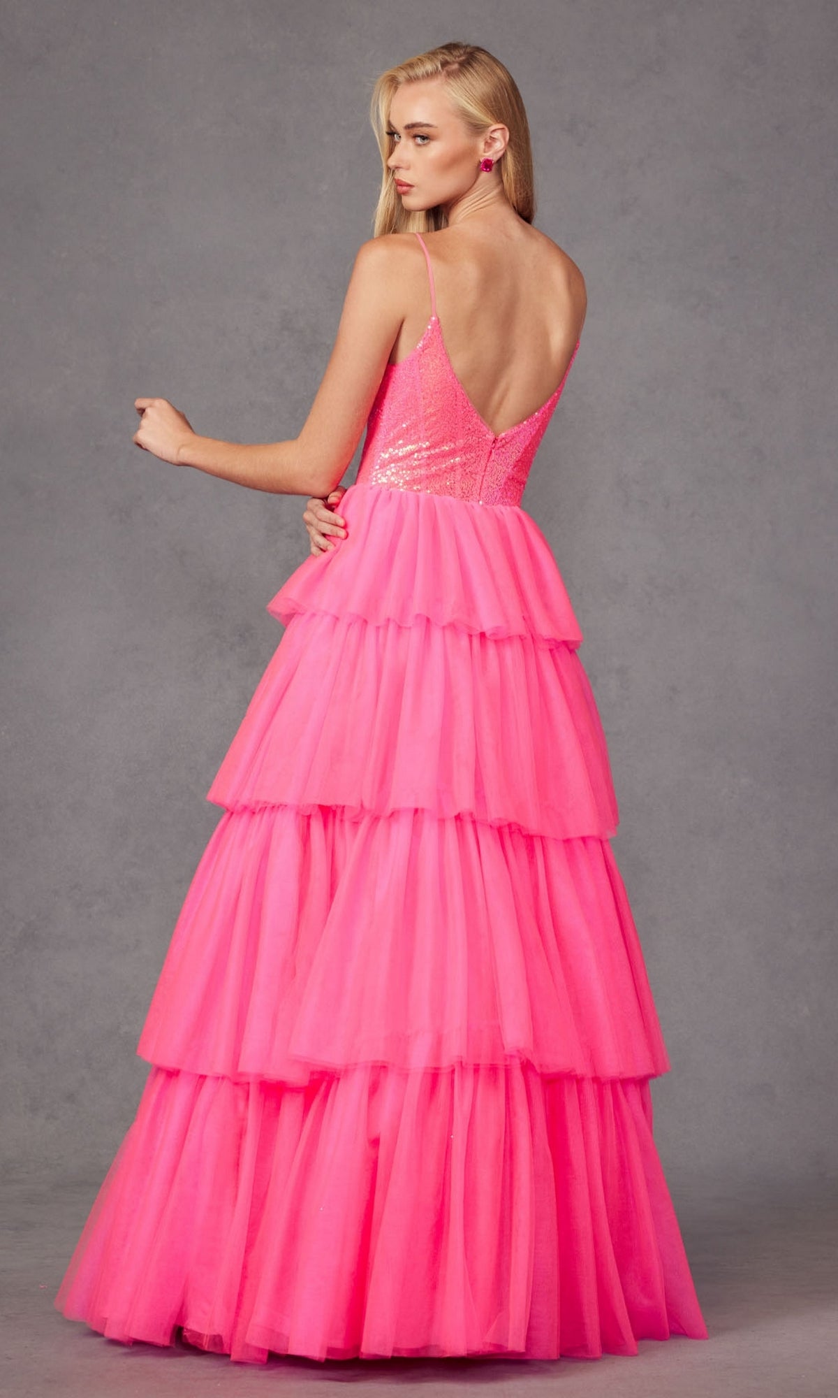 A woman models the Long Prom Dress JT2457H by Juliet—a fuchsia ruffle tulle ball gown with spaghetti straps and a sequin bodice—standing against a gray background, facing away with her head turned to the side.